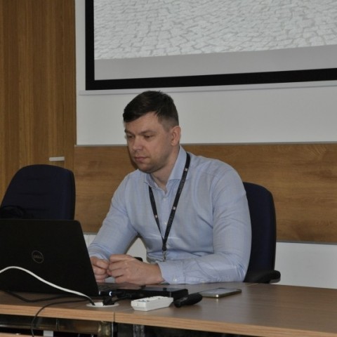 Powiększ obraz: wykładowca, młody mężczyzna w niebieskiej koszuli siedzi